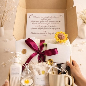 May include: A gift box filled with self-care items. Includes a white lotion bottle, a candle, a mug, a towel, and a card with text. A burgundy ribbon and a yellow crochet sunflower add decorative touches.