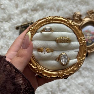 May include: A collection of gold rings displayed in a round, ornate gold frame with a cream-colored interior. The rings feature various designs, including a rectangular stone, a band of small stones, and a marquise-cut stone.