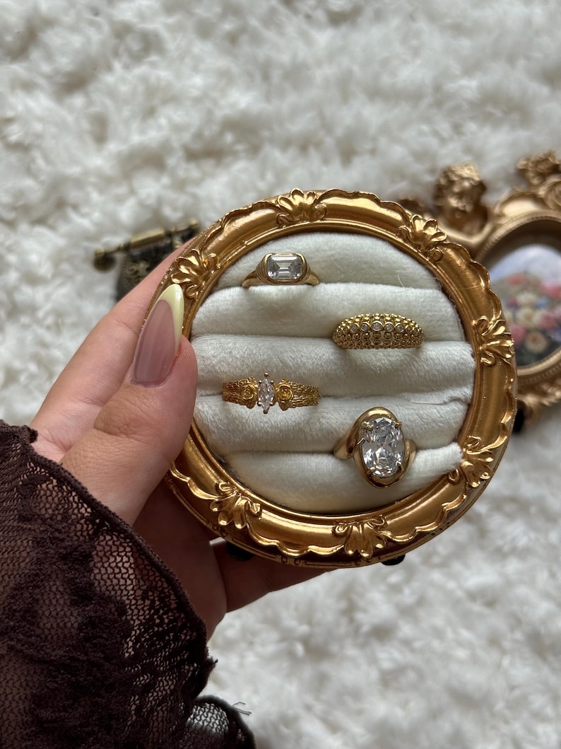 May include: A collection of gold rings displayed in a round, ornate gold frame with a cream-colored interior. The rings feature various designs, including a rectangular stone, a band of small stones, and a marquise-cut stone.