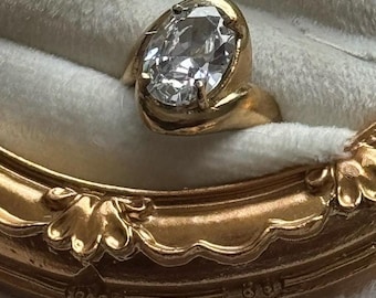 Könnte beinhalten: Ein Goldring mit einem großen, oval geschliffenen, klaren Edelstein, der in eine glatte, polierte Goldschiene gefasst ist. Der Ring befindet sich in einem mit weißem Samt ausgekleideten Etui mit einem verzierten, goldfarbenen Rahmen.