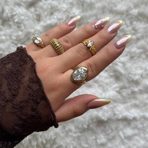 May include: A hand displaying several gold rings with various designs, including a rectangular stone, a textured band, a floral design with a clear stone, and an oval-shaped stone. The nails are long and painted with a light pink and yellow color.
