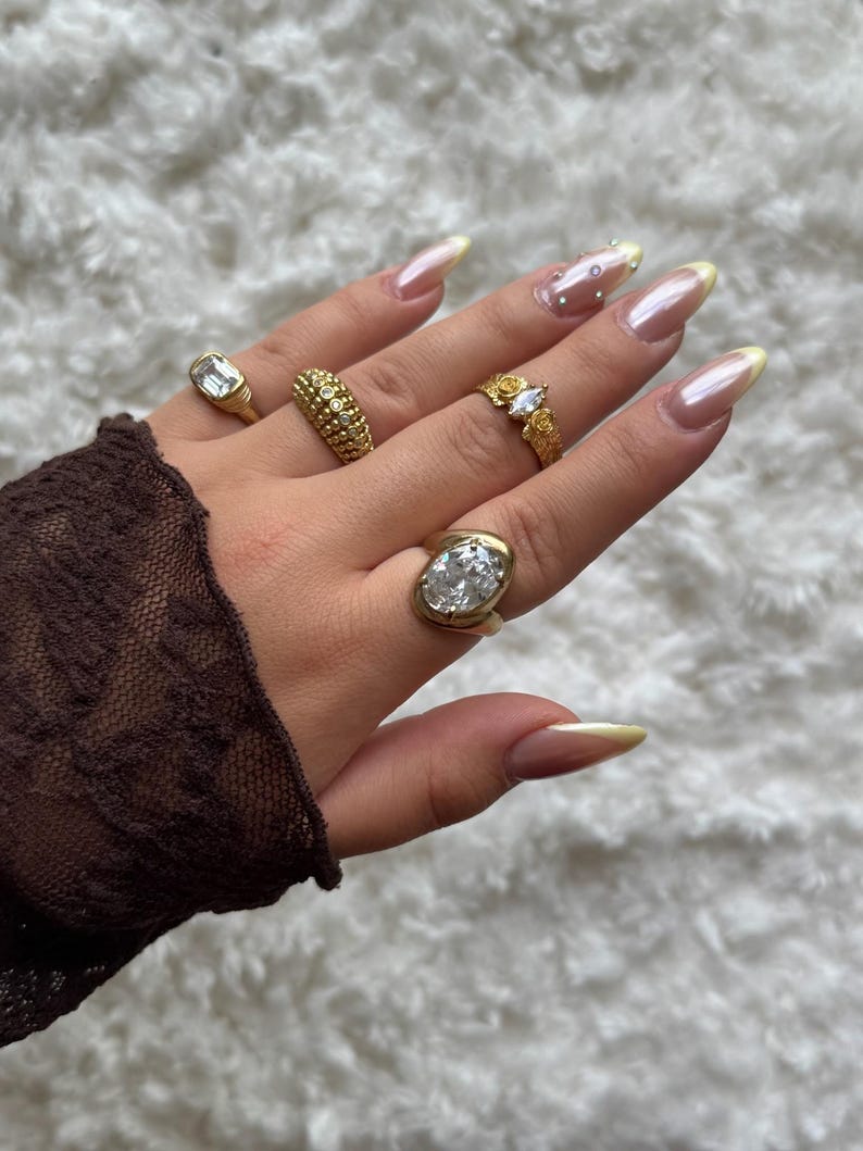 May include: A hand displaying several gold rings with various designs, including a rectangular stone, a textured band, a floral design with a clear stone, and an oval-shaped stone. The nails are long and painted with a light pink and yellow color.