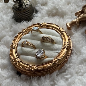 May include: A collection of gold rings with various gemstone settings displayed in a decorative, ornate gold frame. The rings include a rectangular, oval, and round cut stones. The frame sits on a fluffy white surface, with a vintage telephone and cherub statue in the background.