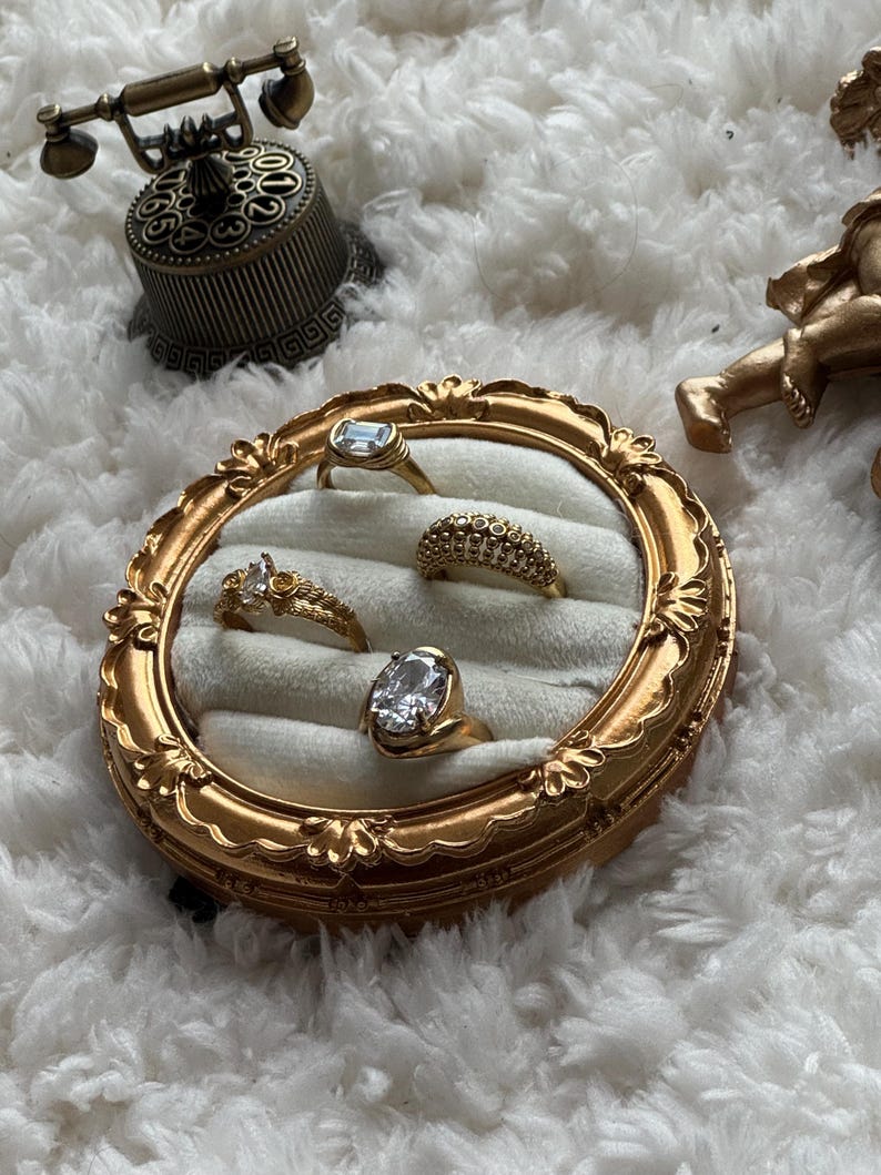 May include: A collection of gold rings with various gemstone settings displayed in a decorative, ornate gold frame. The rings include a rectangular, oval, and round cut stones. The frame sits on a fluffy white surface, with a vintage telephone and cherub statue in the background.