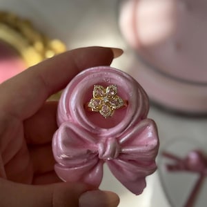 May include: A gold ring featuring a flower-shaped centerpiece with five marquise-cut light pink stones and a small clear center stone, set within a light pink, sculpted, bow-shaped display stand with a glossy, smooth finish.