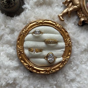 May include: A gold-toned ring display with five rings. The rings feature various designs, including a rectangular stone, a band of small stones, and an oval-shaped stone. The display is set against a white, fluffy background.