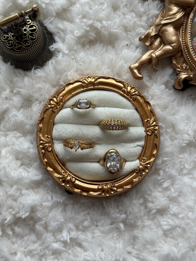 May include: A gold-toned ring display with five rings. The rings feature various designs, including a rectangular stone, a band of small stones, and an oval-shaped stone. The display is set against a white, fluffy background.