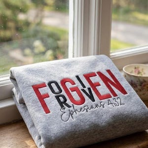 Peut inclure: Un sweat-shirt gris plié avec le mot "FORGIVEN" brodé en rouge et noir, avec "Éphésiens 4:32" en dessous. Une tasse à thé fleurie est posée à côté du sweat-shirt sur un rebord de fenêtre en bois, avec une fenêtre et une vue extérieure en arrière-plan.