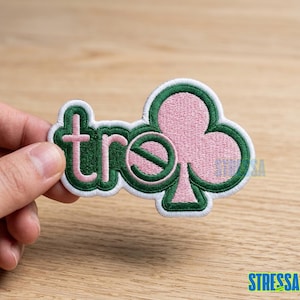 May include: Embroidered patch in the shape of a clover with the word "tre" in pink and green. The patch is held by a hand against a light brown background. The patch has a white border.