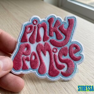 May include: Embroidered patch with the words "Pinky Promise" in a retro font. The text is in a vibrant pink color, outlined in a light blue, and set against a white background. The patch has a light blue border.