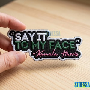 May include: Embroidered patch with the text "SAY IT TO MY FACE" in white and green, with "-Kamala Harris" in pink. The patch has a black background and white border. The patch is held by a hand.