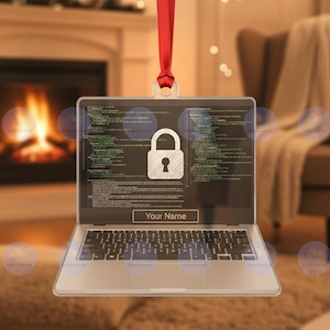 May include: A laptop-shaped ornament with a red ribbon for hanging. The screen displays a padlock icon and lines of code, with the text "Your Name" below. The keyboard is visible, and the background shows a fireplace.