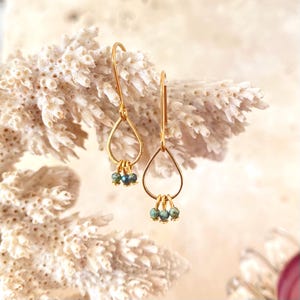 Peut inclure: Boucles d'oreilles dorées en forme de goutte d'eau, ornées de trois petites perles bleu-vert. Elles sont présentées sur un fond blanc texturé, créant un contraste de couleurs et de textures.
