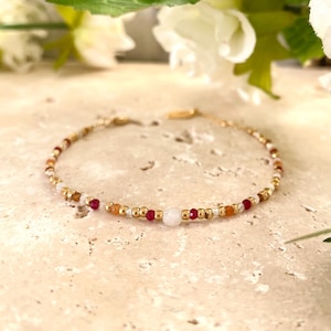 Könnte beinhalten: Zartes goldfarbenes Armband mit einer Mischung aus kleinen, farbigen Perlen. Das Armband enthält rote, orangefarbene, weiße und goldfarbene Perlen. Ein zartes Schmuckstück.