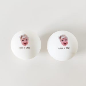 May include: Two white spherical objects featuring a printed photograph of a smiling baby wearing a headband. Below the baby's face, the text reads "Kobie is ONE" in a simple black sans-serif font. The items have a smooth, matte finish.