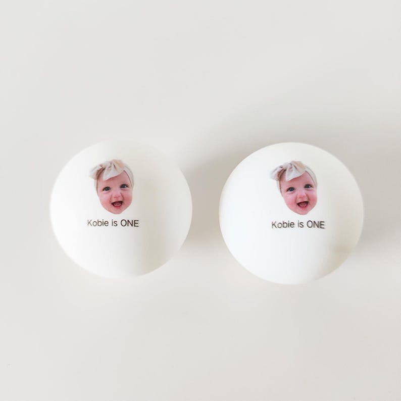 May include: Two white spherical objects featuring a printed photograph of a smiling baby wearing a headband. Below the baby's face, the text reads "Kobie is ONE" in a simple black sans-serif font. The items have a smooth, matte finish.