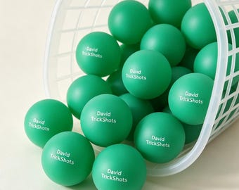 May include: A collection of matte green ping pong balls inside a white plastic grid basket. Each ball features the white printed text David TrickShots. The balls are spherical and uniform in size, spilling out from the open side of the container.