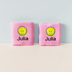 May include: A pair of light pink terry cloth wristbands, each featuring a graphic of a smiling yellow tennis ball and the name Julia printed in black text below the ball. The fabric has a soft, textured appearance consistent with athletic gear.