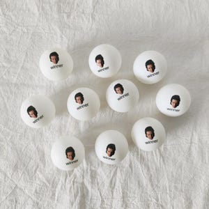 May include: A collection of nine white ping pong balls, each featuring a small printed portrait of a person and the word winner in black text. The balls are arranged in a loose cluster, showcasing the identical graphic design on each surface.