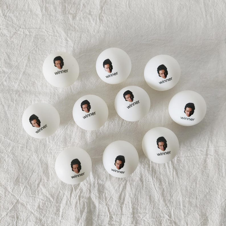 May include: A collection of nine white ping pong balls, each featuring a small printed portrait of a person and the word winner in black text. The balls are arranged in a loose cluster, showcasing the identical graphic design on each surface.