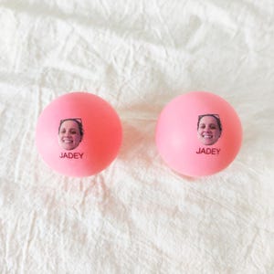 May include: Two round, bright pink stress balls, each featuring a small, centered photographic print of a smiling person's face above the printed name JADEY in black capital letters. The surface of the balls is smooth and matte.