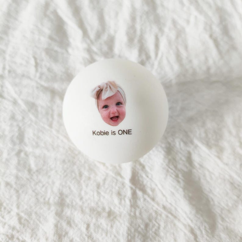 May include: A white spherical ball featuring a printed photograph of a smiling baby wearing a bow headband. Below the image, the text reads Kobie is ONE in black lettering. The object has a smooth, matte surface finish.