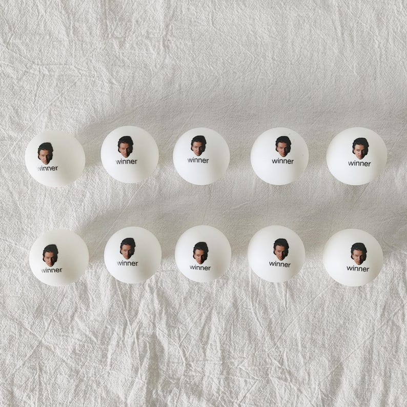 May include: Ten white ping pong balls arranged in two rows of five. Each ball features a small black and white portrait of a person's face above the word winner printed in black lowercase letters on the surface of the ball.