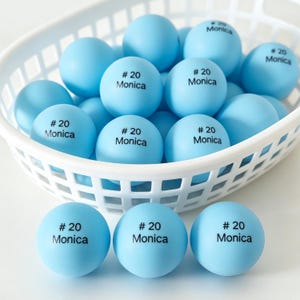 Könnte beinhalten: Eine Sammlung hellblauer Tischtennisbälle in einem weißen Kunststoffkorb. Jeder Ball ist mit dem schwarzen Text # 20 Monica bedruckt. Die Bälle sind glatt und kugelförmig, wobei mehrere im Korb angeordnet sind und drei davor liegen.