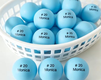 May include: A collection of light blue ping pong balls inside a white plastic basket. Each ball features the black printed text # 20 Monica. Three additional balls with the same text are positioned in front of the basket on a flat surface.