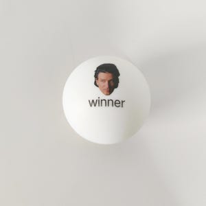 May include: A smooth white spherical object featuring a small, centered photographic portrait of a person with dark hair above the lowercase black text that reads winner. The object has a clean, matte finish and a simple, rounded design.