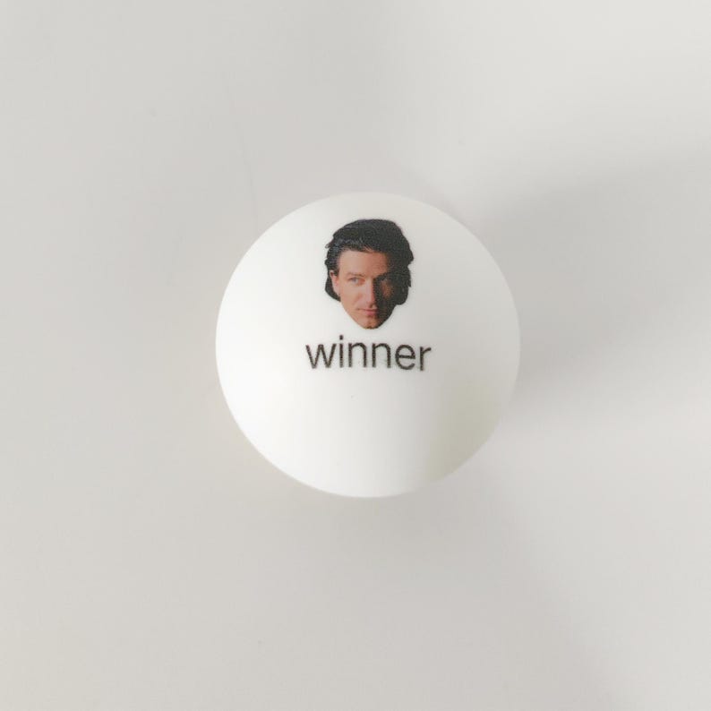 May include: A smooth white spherical object featuring a small, centered photographic portrait of a person with dark hair above the lowercase black text that reads winner. The object has a clean, matte finish and a simple, rounded design.