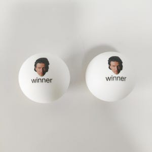 May include: Two white spherical ping pong balls, each featuring a small color photograph of a person's face printed above the lowercase black text "winner" centered on the front surface of the ball.