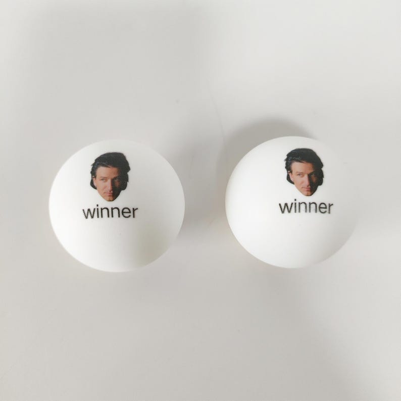 May include: Two white spherical ping pong balls, each featuring a small color photograph of a person's face printed above the lowercase black text "winner" centered on the front surface of the ball.
