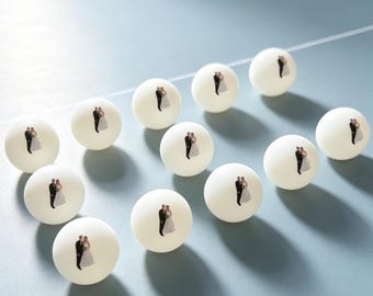 May include: A collection of twelve white ping pong balls, each featuring a small, printed graphic of a bride and groom standing together. The balls are arranged in a grid-like pattern, showcasing the identical wedding-themed imagery on each unit.