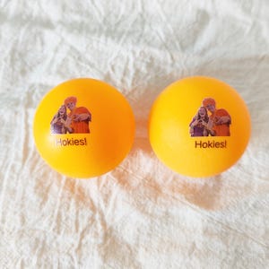 May include: Two bright orange ping pong balls, each featuring a small printed graphic of two people making hand gestures and the text Hokies! printed in a dark color below the image on the smooth, spherical surface of the balls.