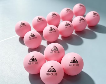 May include: A collection of fifteen light pink ping pong balls arranged in a cluster. Each ball features a black printed logo consisting of a stylized triangular icon above the text GB CLUB. The balls have a smooth, matte finish and round shape.