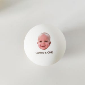 May include: A round white ball featuring a printed photograph of a smiling baby's face in the center. Below the baby's face, the text "Caffrey is ONE" is printed in a simple black sans-serif font on the smooth white surface.