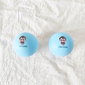 May include: Two light blue spherical balls, each featuring a small photographic print of a person's face and the text CON CONS printed in black ink below the image. The balls have a smooth, matte surface and are identical in appearance.