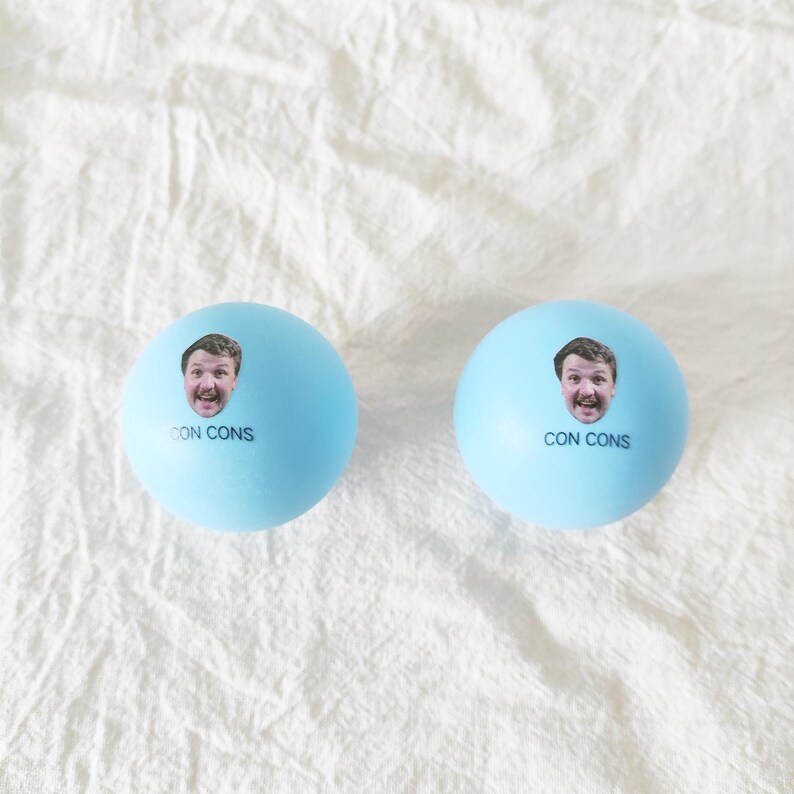 May include: Two light blue spherical balls, each featuring a small photographic print of a person's face and the text CON CONS printed in black ink below the image. The balls have a smooth, matte surface and are identical in appearance.