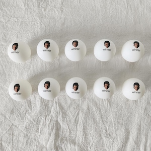 May include: Ten white ping pong balls arranged in two rows of five. Each ball features a small printed graphic of a person's face above the word winner in black lowercase letters. The balls have a smooth matte finish and spherical shape.