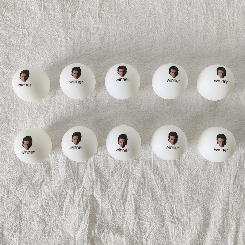 May include: Ten white ping pong balls arranged in two rows of five. Each ball features a small printed graphic of a person's face above the word winner in black lowercase letters. The balls have a smooth matte finish and spherical shape.