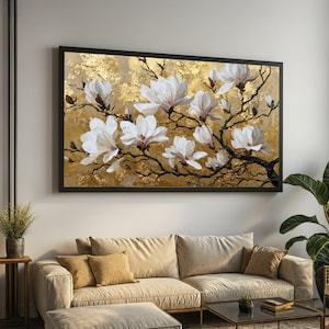 以下が含まれることがあります： 金色のテクスチャ背景に白いモクレンの花を描いた額入りの絵画。 黒いフレームに入ったこのアートワークは、ベージュのソファの上に飾られています。 絵画の花柄は、インテリアに自然な要素を加えています。