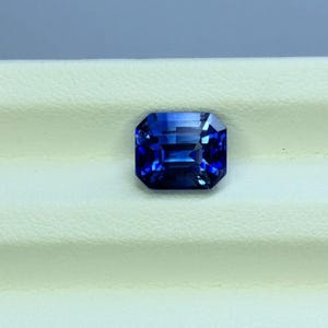 Puede incluir: Una piedra preciosa de color azul profundo con corte esmeralda y una superficie facetada que refleja la luz. La piedra presenta una forma rectangular con esquinas recortadas y exhibe un perfil de color rico y saturado con claridad interna.