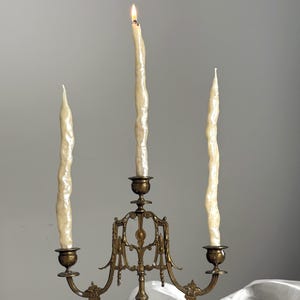 May include: A brass candelabra with three ivory-colored, textured candles. One candle is lit, with a visible flame. The candelabra has an ornate design with curved arms and decorative details, suitable for home decor.