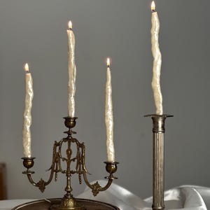 May include: Four lit ivory candles in antique brass candle holders. The candles have a textured, melted appearance. One candle is in a tall, fluted holder. The other three are in a candelabra-style holder with decorative arms.