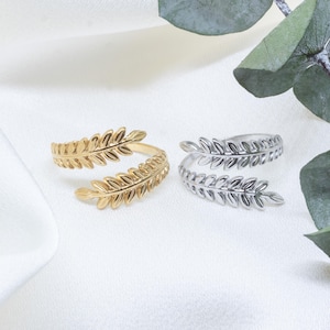 Puede incluir: Dos anillos con forma de hoja. Uno es dorado y el otro plateado. Los anillos están sobre una superficie blanca con una hoja verde de fondo. Los anillos tienen un diseño envolvente.