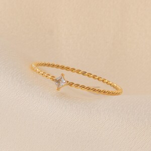May include: A gold-colored ring with a twisted band and a square, clear gemstone. The ring is set against a soft, off-white background, highlighting the jewelry's delicate design and sparkle.