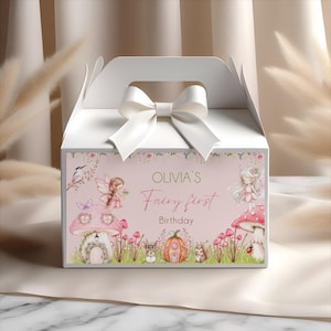 May include: A white gift box with a handle and a white satin bow on top. The front of the box features a pink background with the text "Olivia's Fairy first Birthday" and illustrations of fairies, mushrooms, and woodland creatures.