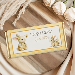 Op de afbeelding: Een rechthoekige chocoladereep met een geel ruitpatroon en gouden folieranden. De reep heeft de tekst "Happy Easter Charlotte" en illustraties van konijntjes en vlinders. Gepresenteerd op een houten serveerplank.