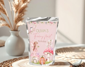 May include: A pink pouch with the text "Olivia's Fairy First Birthday" and illustrations of fairies, mushrooms, and a pumpkin. The pouch is displayed on a wooden slice, with a vase of dried flowers in the background.